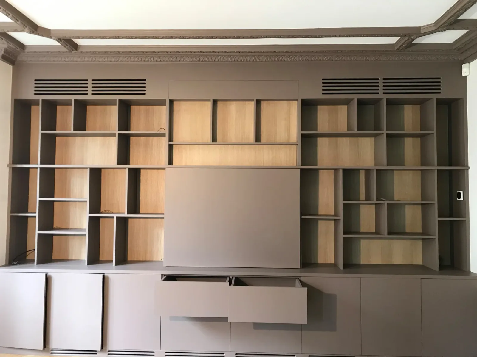 Après travaux : la bibliothèque sur mesure terminée, damier de cases en bois clair et structure taupe, grilles de ventilation intégrées et panneau central dissimulant le téléviseur