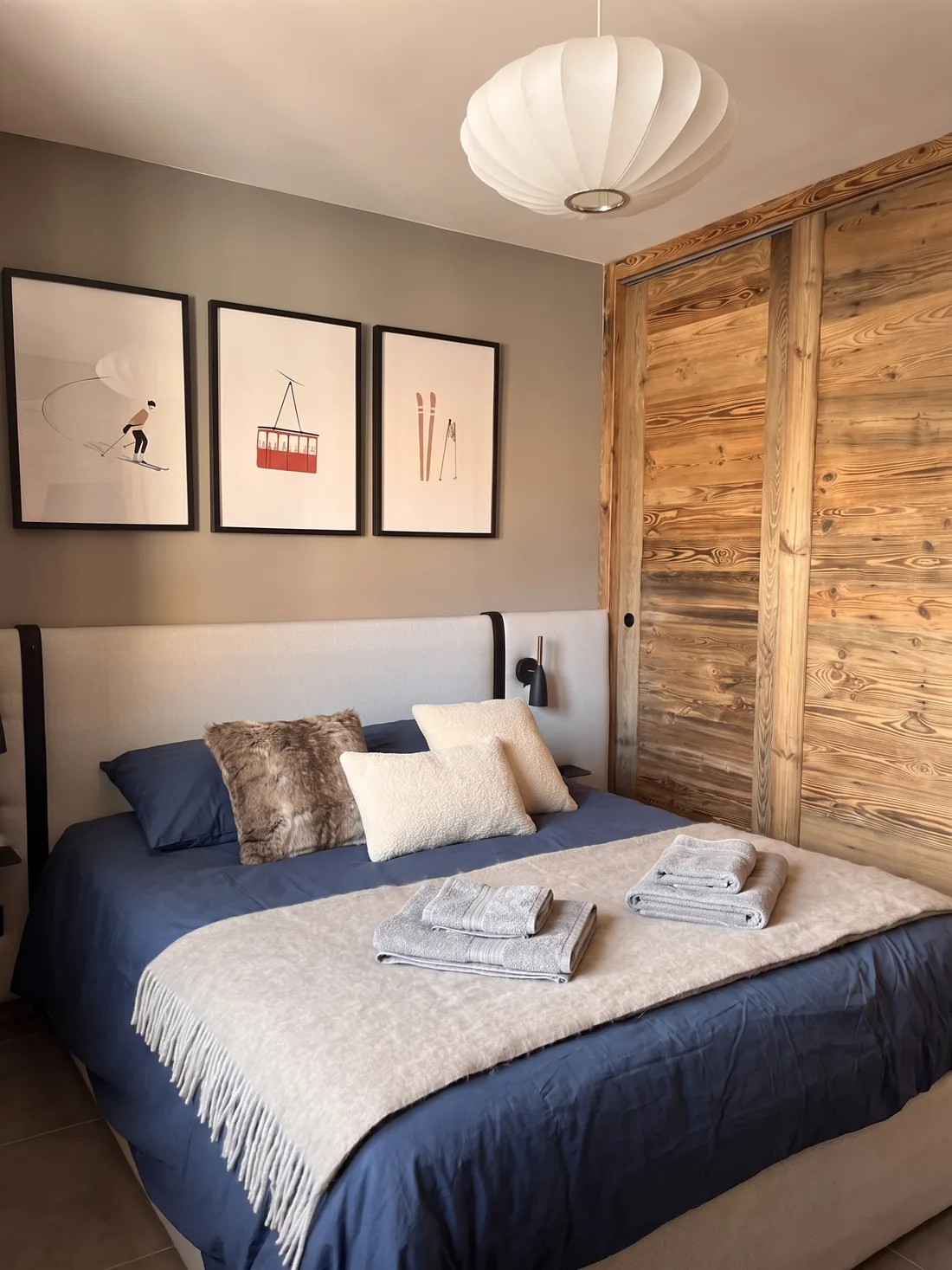 Chambre principale : lit en linge bleu nuit, tête de lit tendue de laine écrue, trois affiches de ski encadrées et grands placards à portes coulissantes en vieux bois