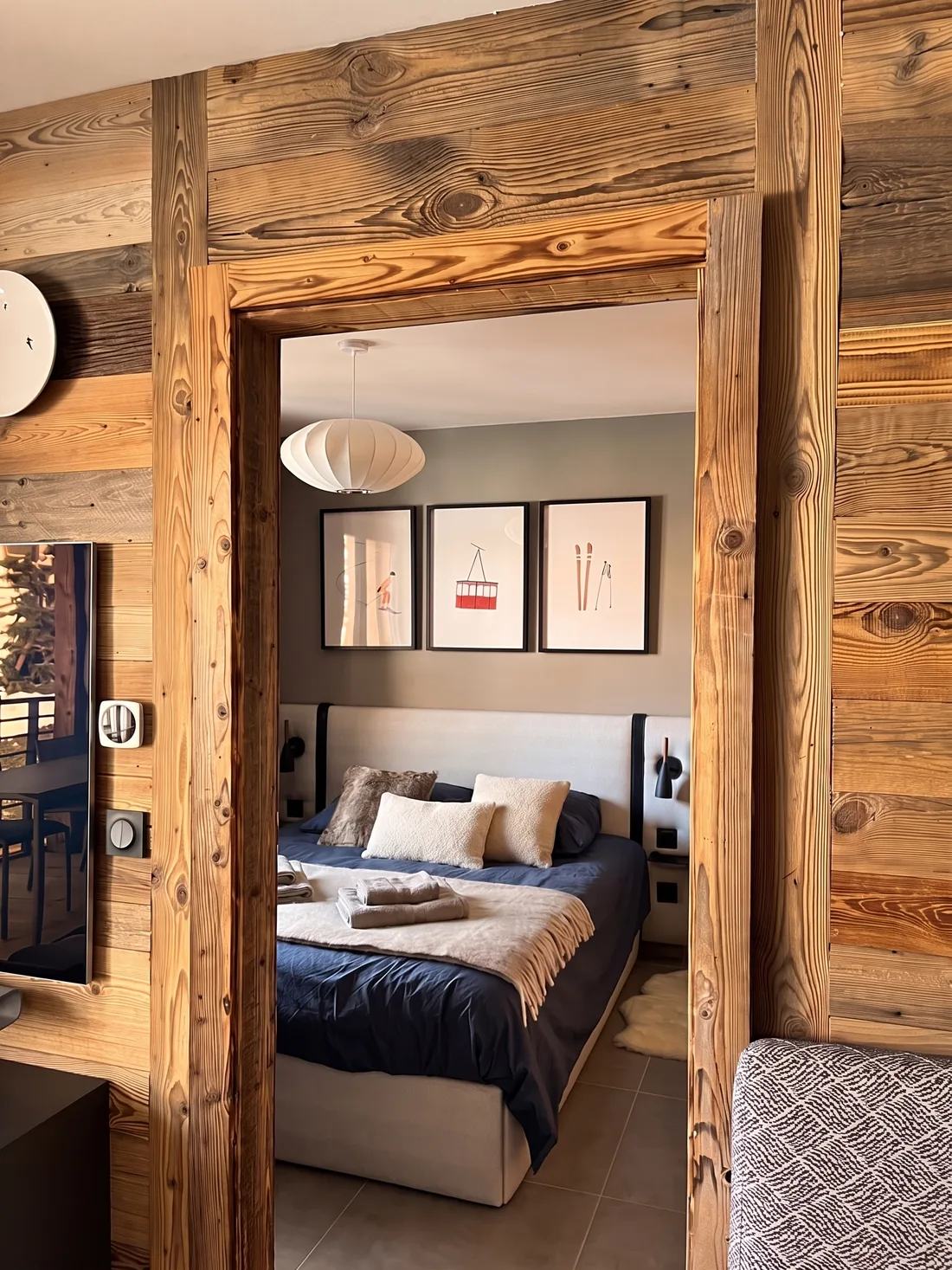 Ouverture encadrée de vieux bois entre le séjour et la chambre : on aperçoit le lit en linge bleu nuit, trois affiches de ski encadrées et une suspension boule blanche plissée