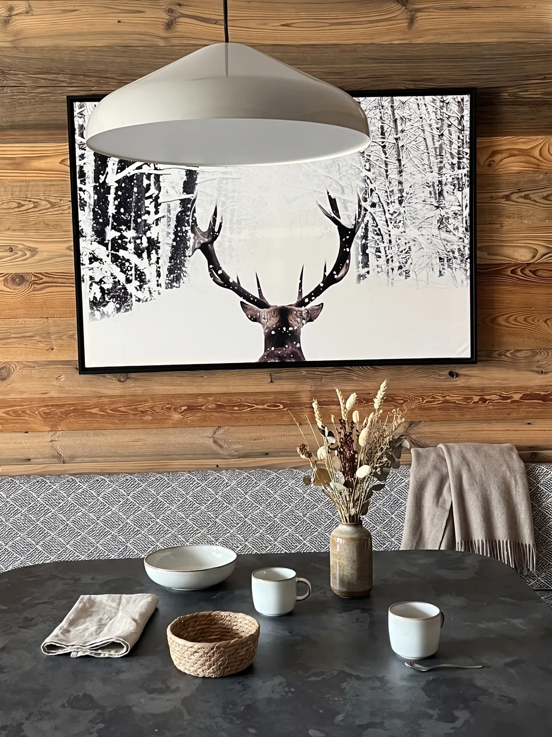Détail du coin repas : grande photographie encadrée d'un cerf dans une forêt enneigée sur le mur de vieux bois, suspension blanche en dôme et plateau de table en pierre bronze