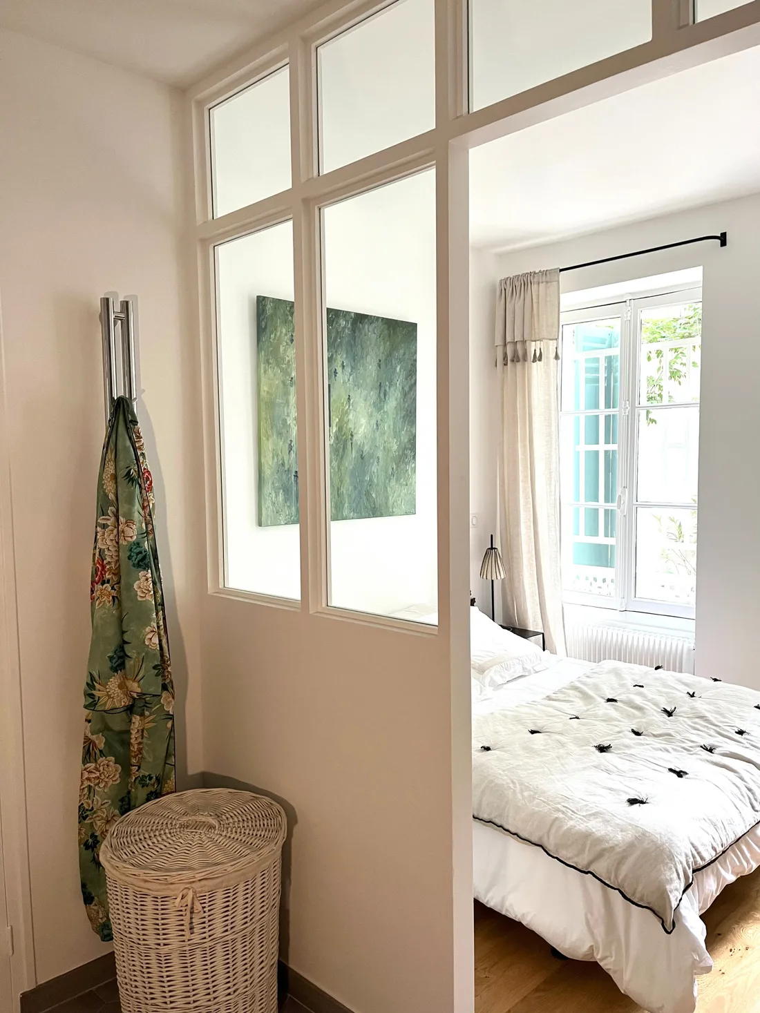La cloison vitrée vue de l'autre côté : impostes blanches, toile abstraite verte en transparence, chambre au couvre-lit à pompons noirs, panier d'osier et kimono fleuri suspendu au sèche-serviettes