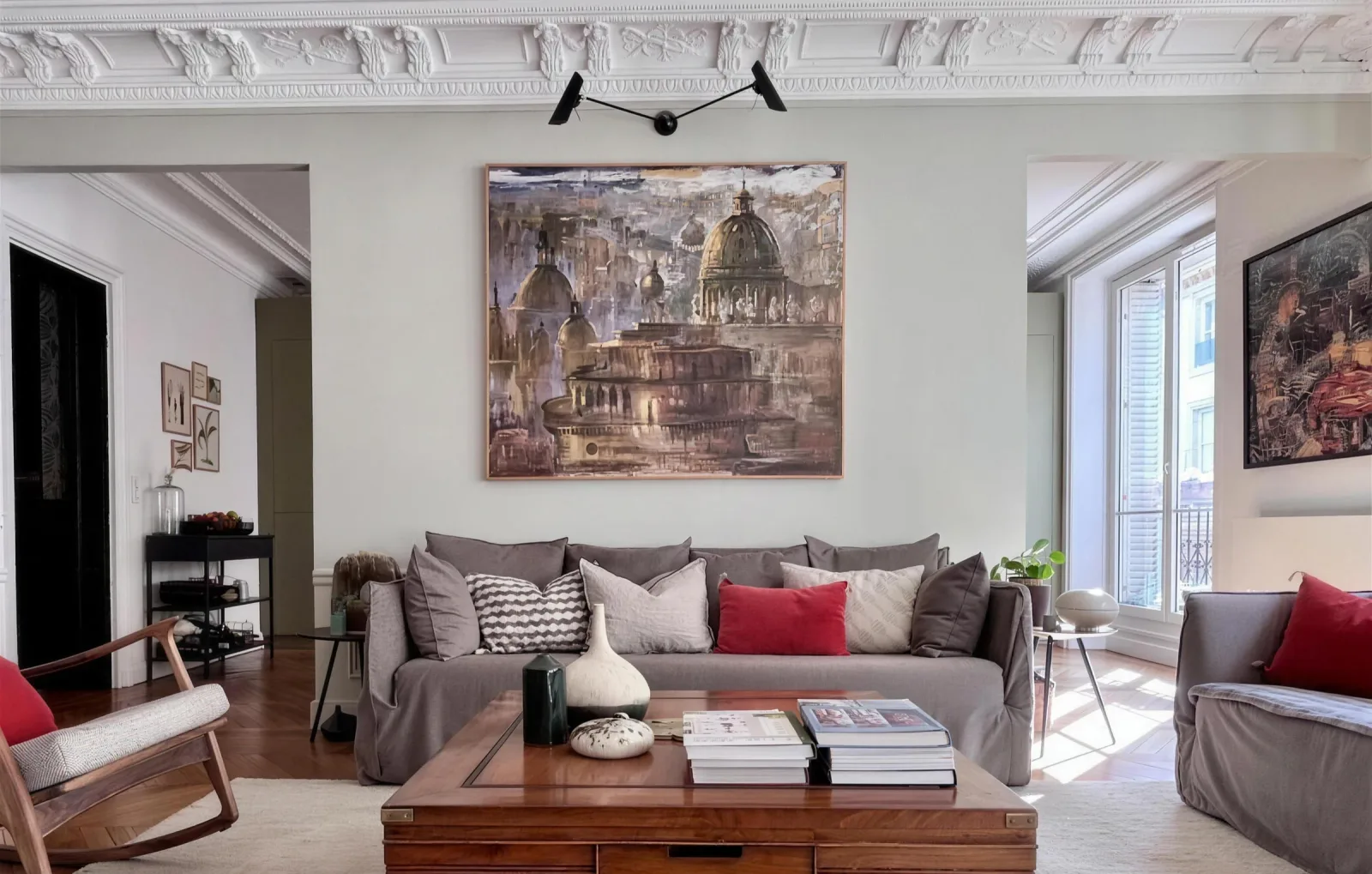 Le salon de l'appartement haussmannien : corniches sculptées, grande toile aux coupoles, applique noire à deux bras, canapé gris aux coussins rouges et table basse en bois