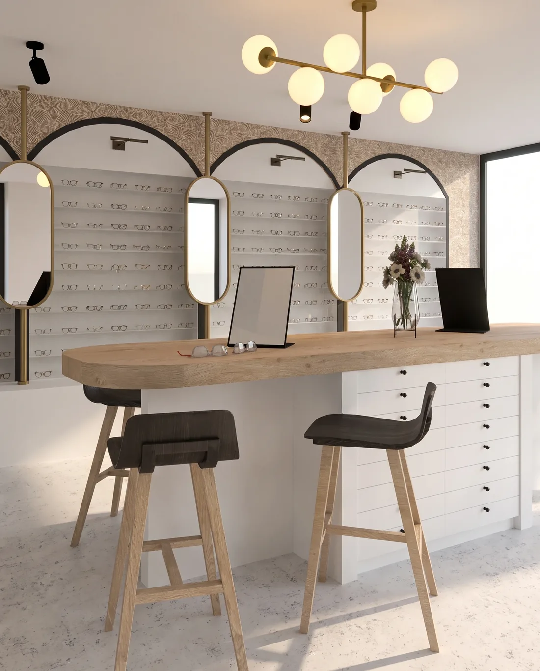 La table d'essayage centrale en bois clair, tabourets à assise noire, miroirs de table, tiroirs blancs à boutons noirs en soubassement et suspension à globes de laiton