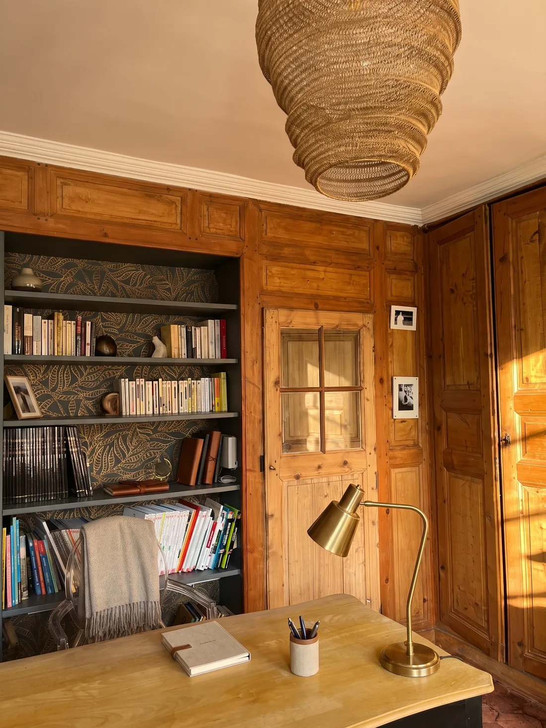 Le bureau aux boiseries de pin ancien : porte vitrée à petits carreaux, bibliothèque encastrée peinte en bleu-gris sur papier peint feuillage doré, suspension de corde tressée et chaise transparente