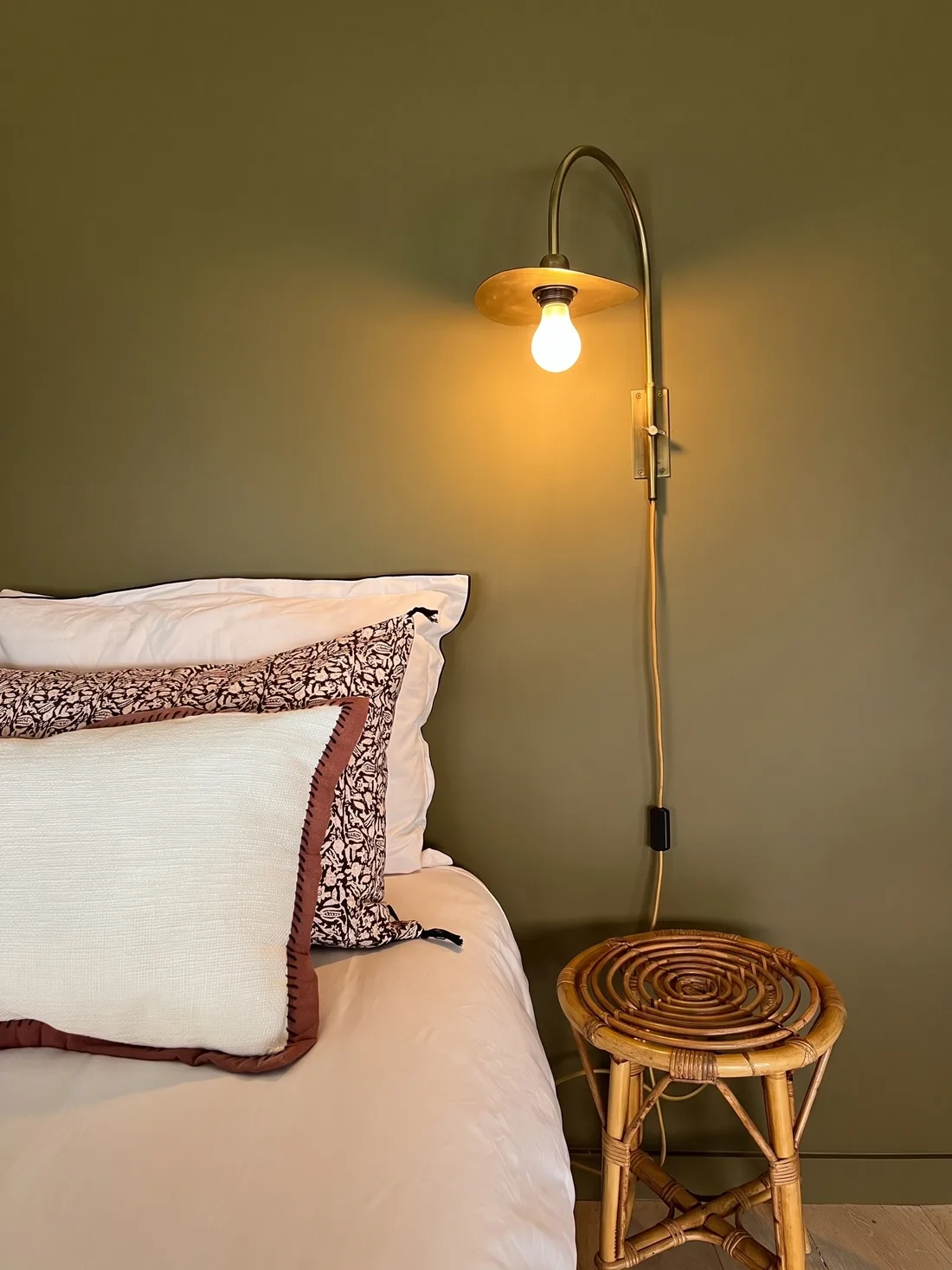 Détail de la chambre principale : mur kaki, liseuse de laiton en col de cygne, oreillers blancs à passepoil fleuri et tabouret de rotin en chevet