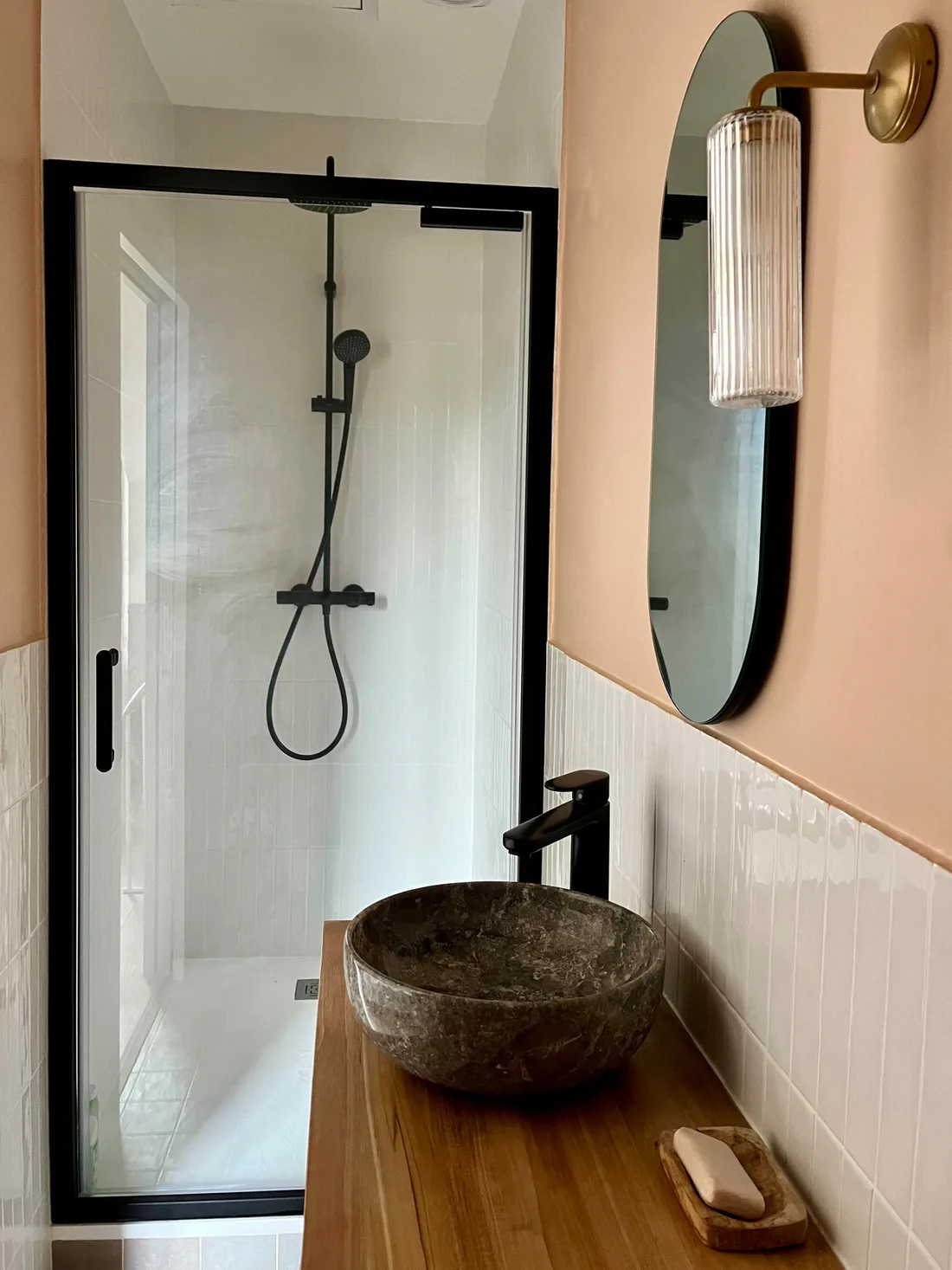 La même salle d'eau côté douche : parois vitrées à châssis noir, zellige blanc vertical, murs rose pêche, vasque ronde de pierre grise sur plan de teck et applique de laiton à verre strié
