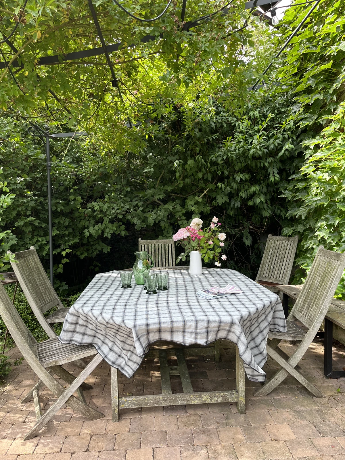 La table du jardin sous la pergola de métal noir : nappe à carreaux, chaises en bois patiné, bouquet de pivoines