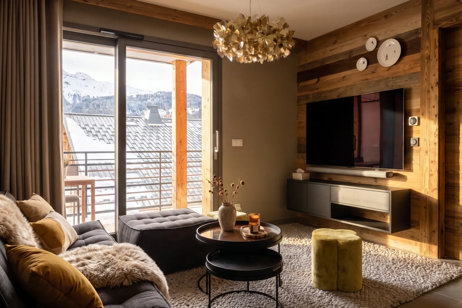 Séjour d'un appartement aux Gets, en Haute-Savoie : mur télé en vieux bois, suspension dorée et montagne enneigée visible par la baie vitrée