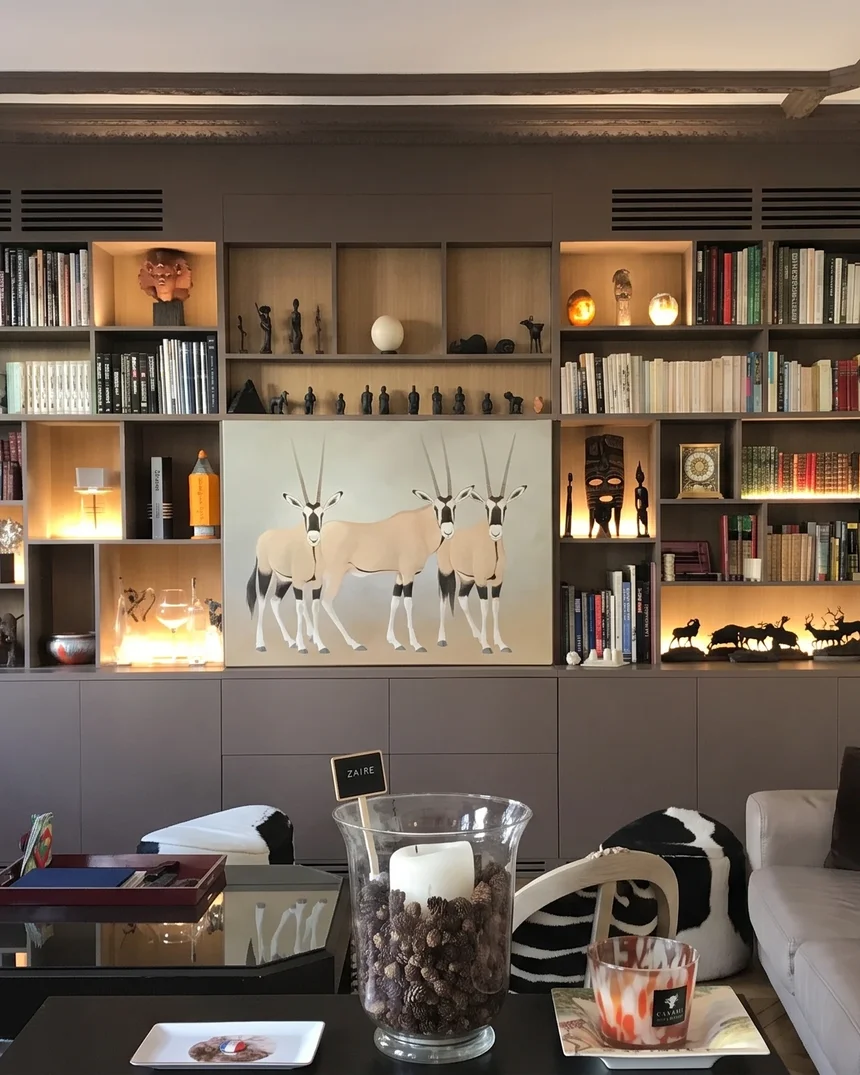 La bibliothèque sur mesure taupe, éclairée niche par niche : livres, statuettes africaines, toile aux oryx masquant l'écran et trophée d'oryx au mur