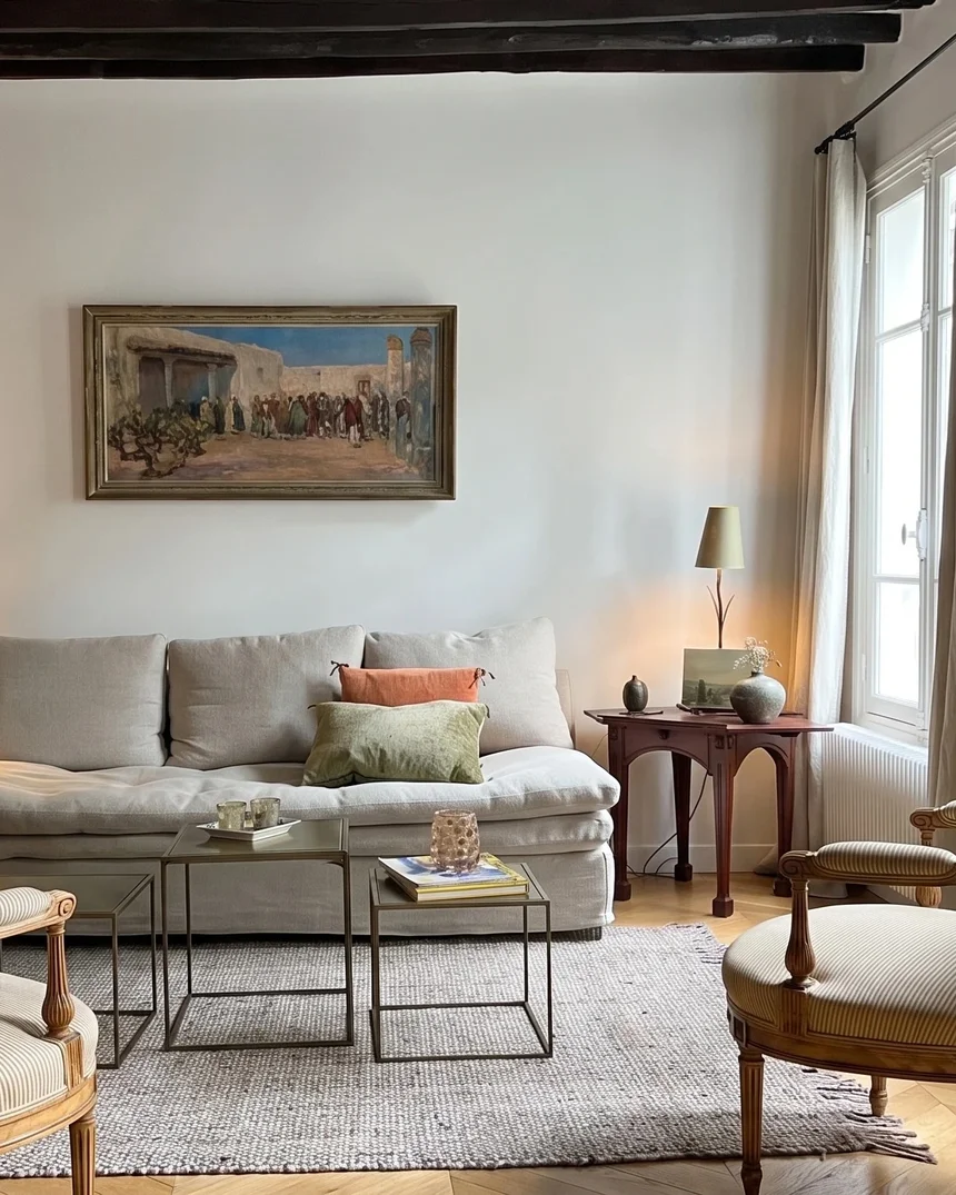 Le salon du duplex ancien : plafond à poutres sombres, canapé de lin écru sous un tableau orientaliste, deux fauteuils Louis XVI à rayures et tables gigognes de métal
