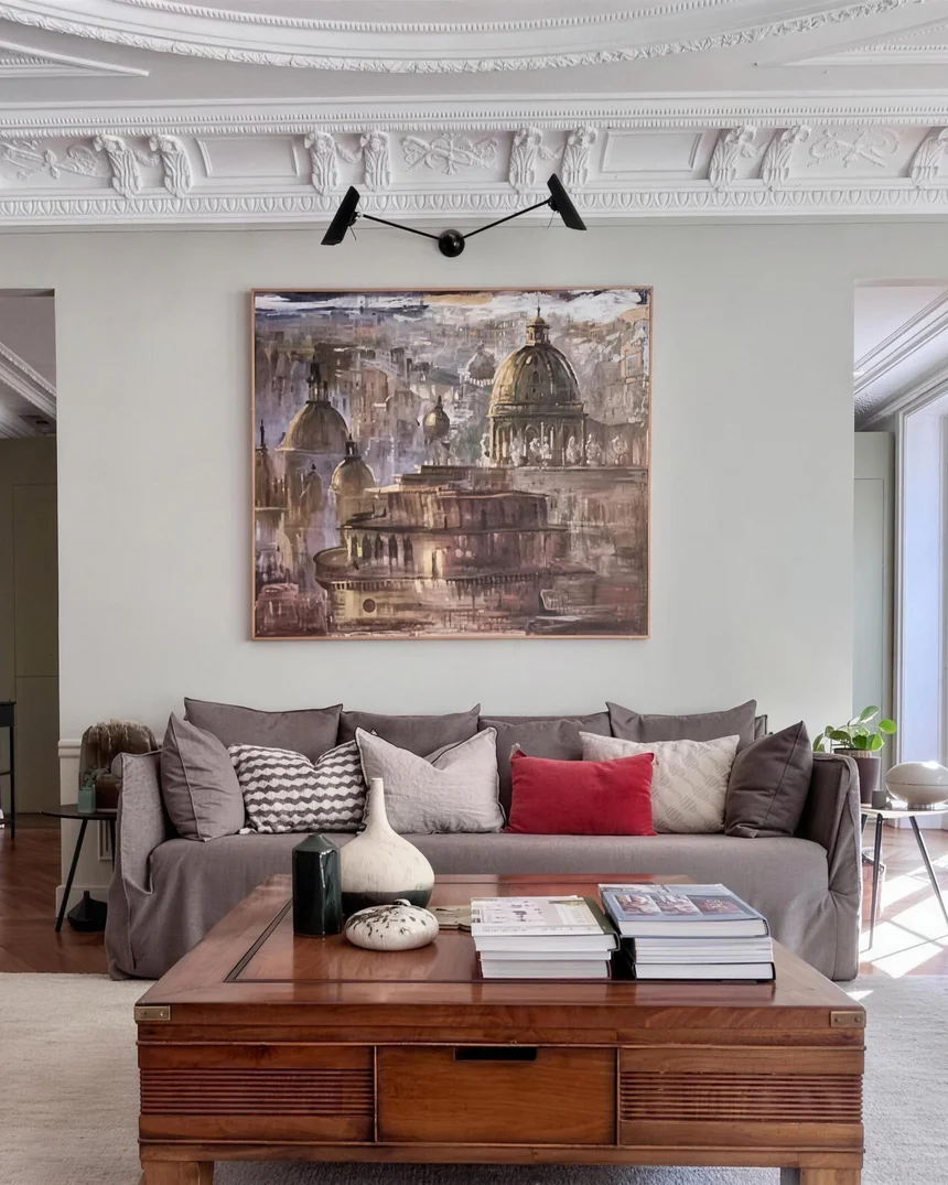 Salon haussmannien décoré : corniches sculptées, applique noire à deux bras, grande toile aux dômes dorés au-dessus d'un canapé gris aux coussins écrus et rouge, parquet point de Hongrie