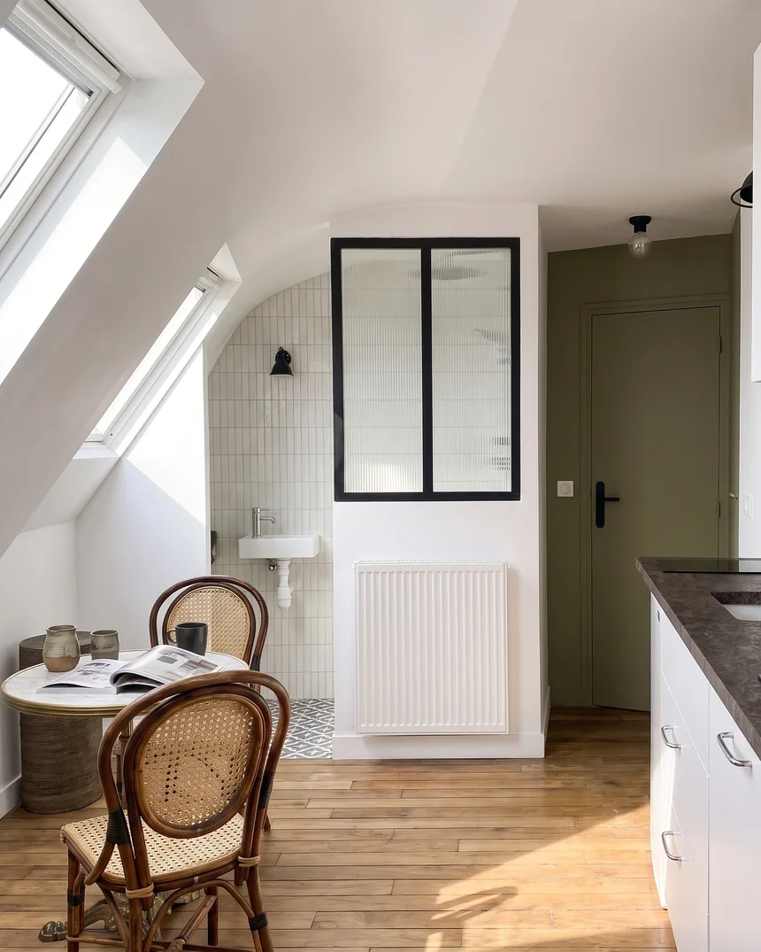 Le studio sous les toits : deux fenêtres de toit, coin repas bistrot en rotin, verrière noire de la salle d'eau carrelée de blanc et porte vert olive