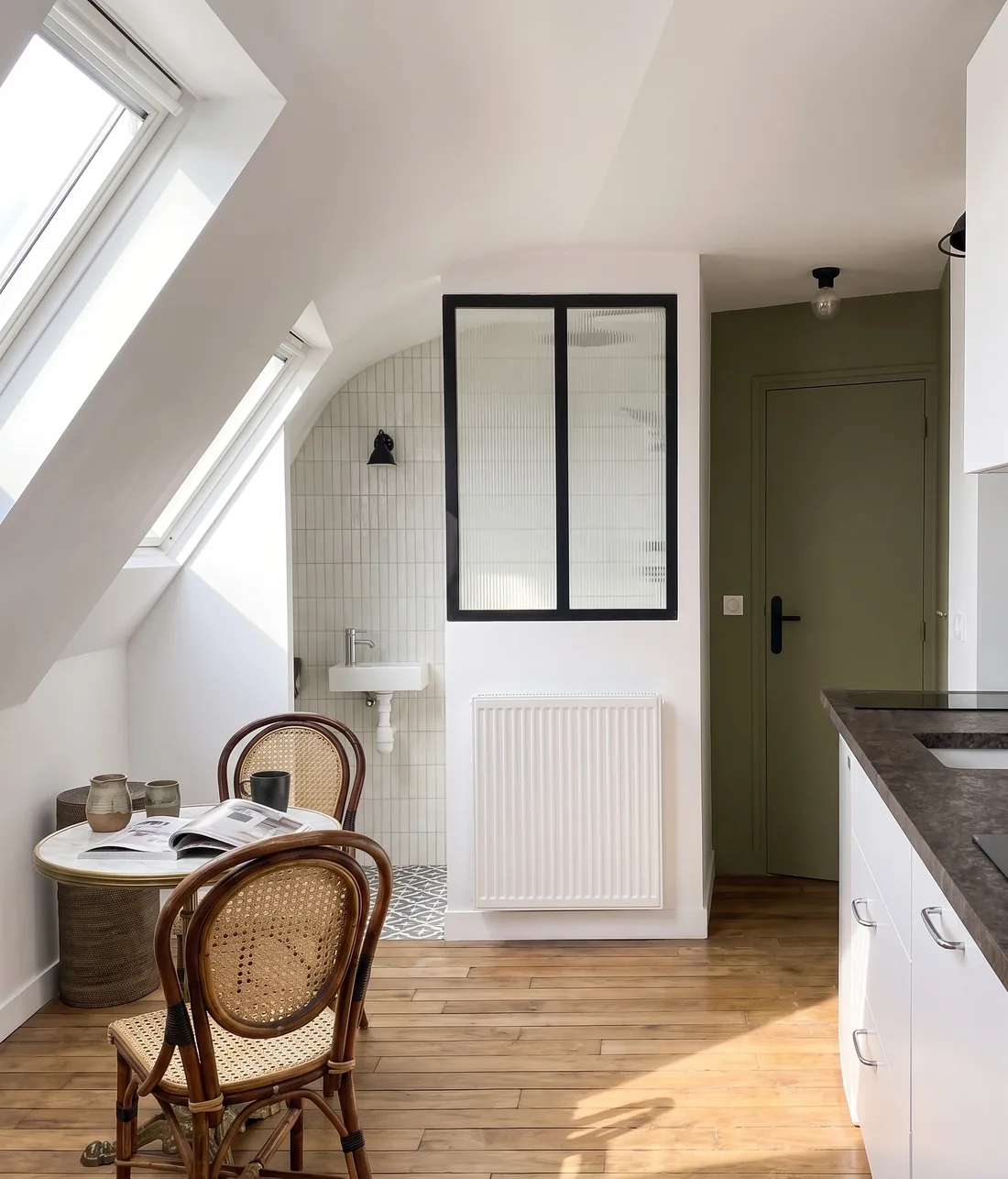 La salle d'eau derrière sa verrière à châssis noir, lavabo et miroir éclairés, radiateur sous la cloison et kitchenette à plan sombre au premier plan