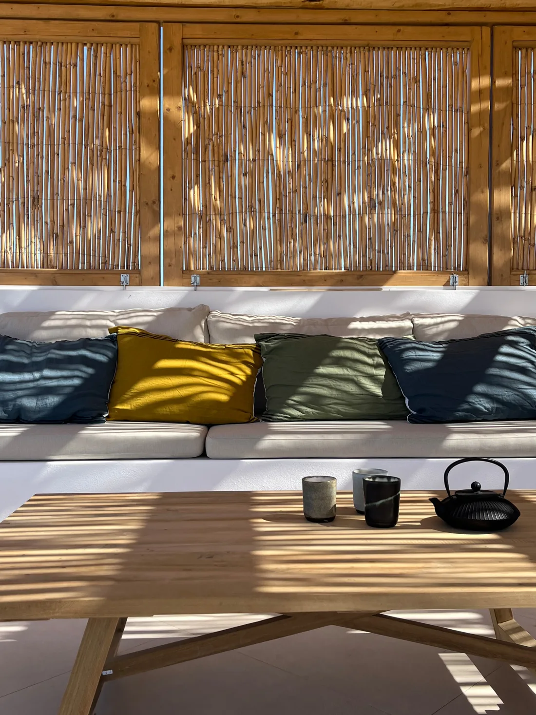 Détail de la banquette sous les claustras de bambou : coussins de lin bleu, moutarde et kaki, table basse de bois clair, théière de fonte noire et lumière rayée du soir