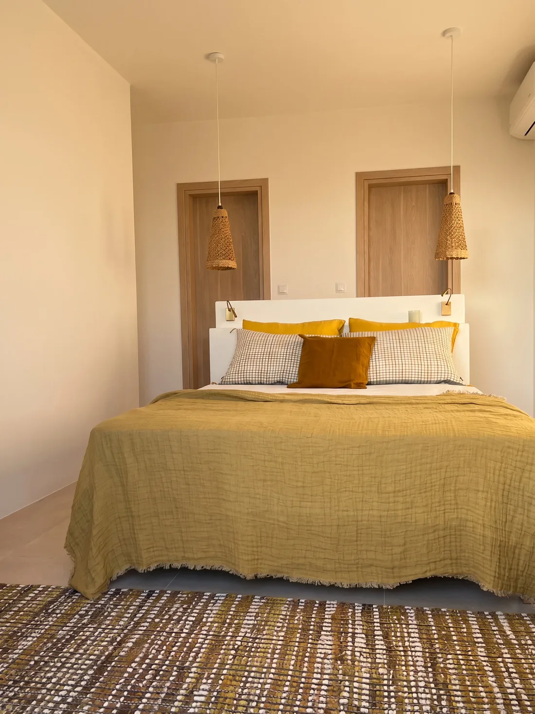 Chambre aux gazes jaune et olive : tête de lit maçonnée blanche, liseuses en laiton encastrées, deux suspensions de raphia tressé et tapis tissé multicolore