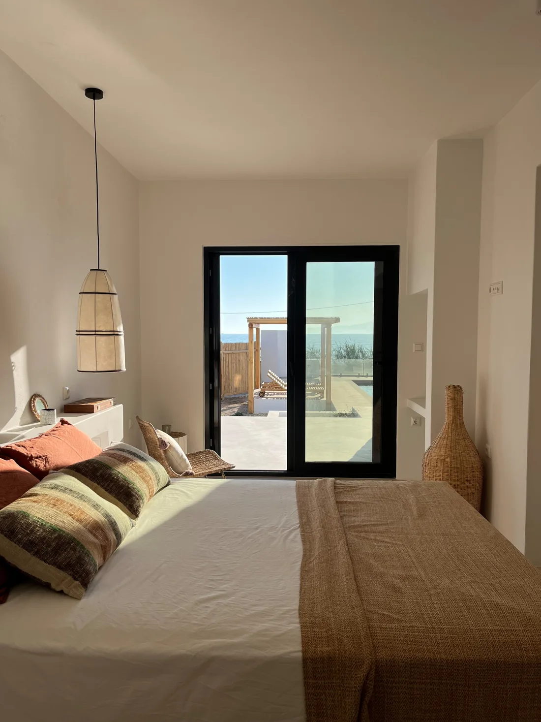 Chambre ouvrant sur la terrasse par une porte vitrée à châssis noir : lit au plaid tissé, coussins terracotta et rayés, dame-jeanne d'osier et mer en toile de fond