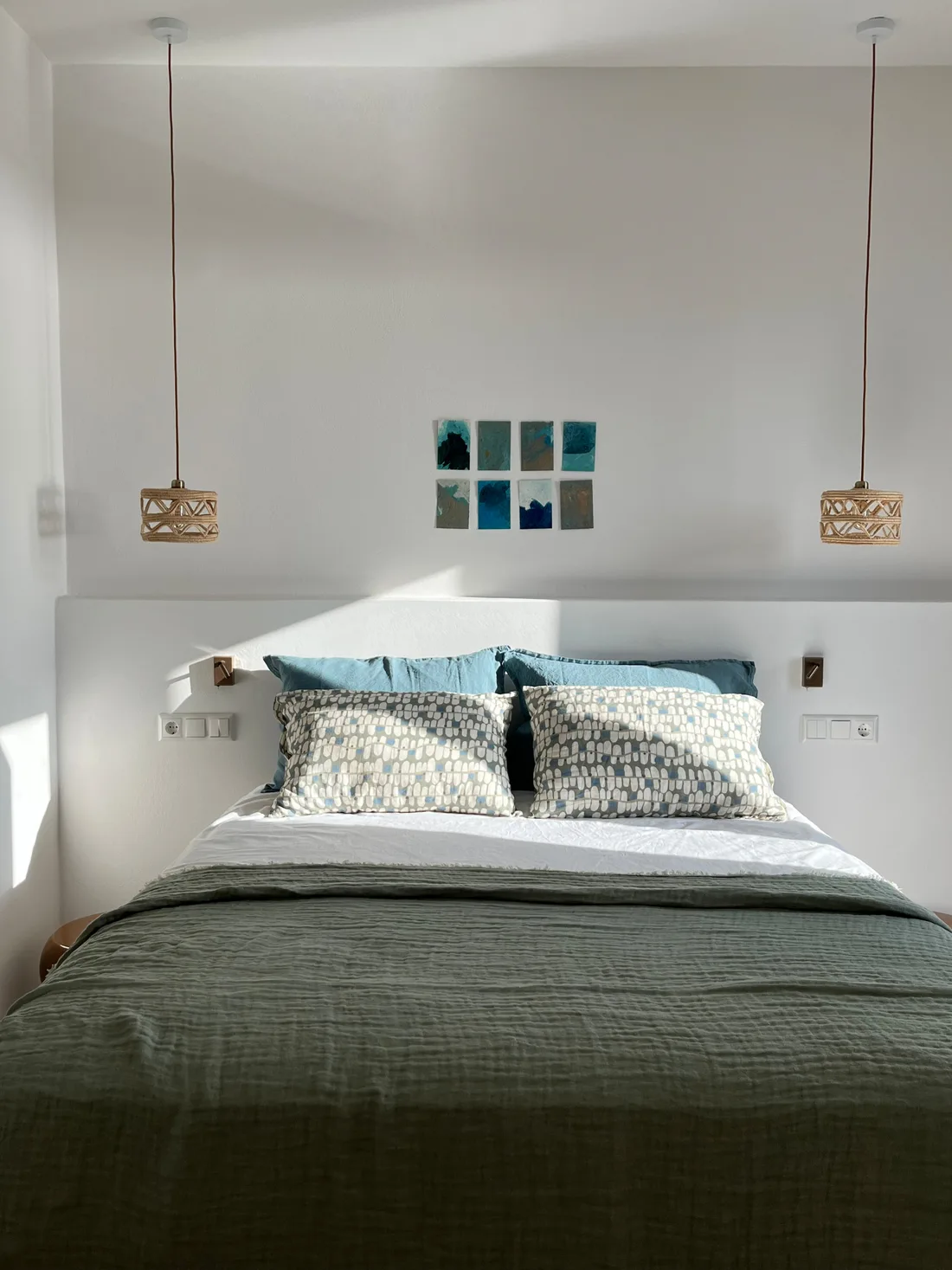 Chambre au couvre-lit kaki : tête de lit maçonnée, coussins bleus, deux suspensions de raphia et grille de huit petites toiles abstraites bleues au mur