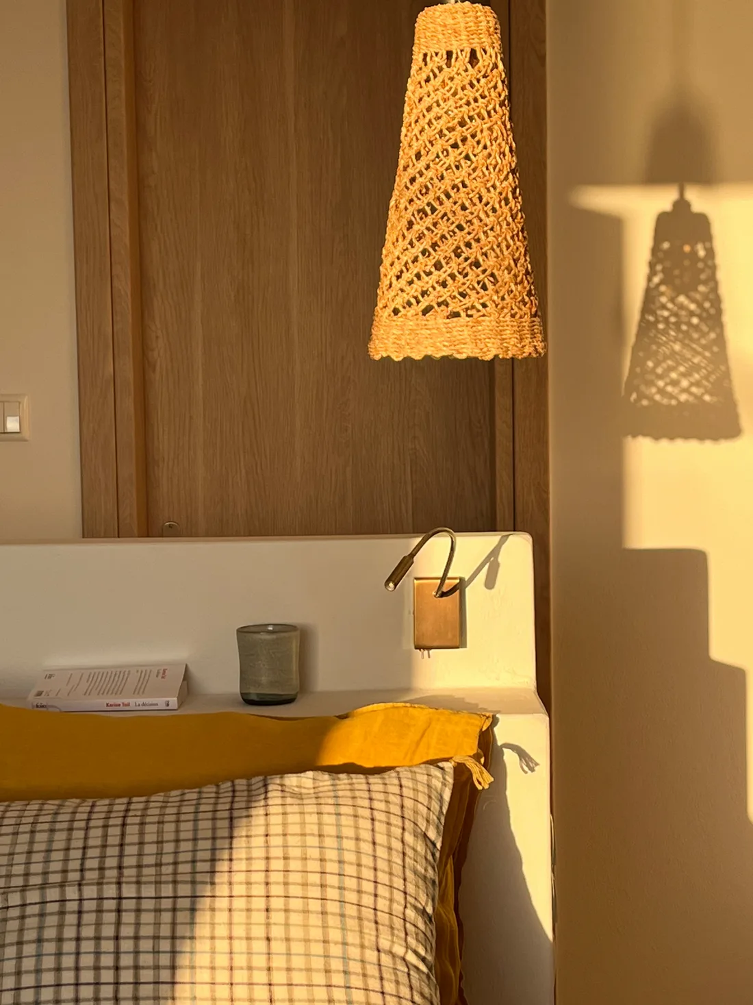 Détail d'un chevet au soir : suspension de raphia projetant son ombre sur le mur, liseuse de laiton, tête de lit maçonnée et linge de lit moutarde