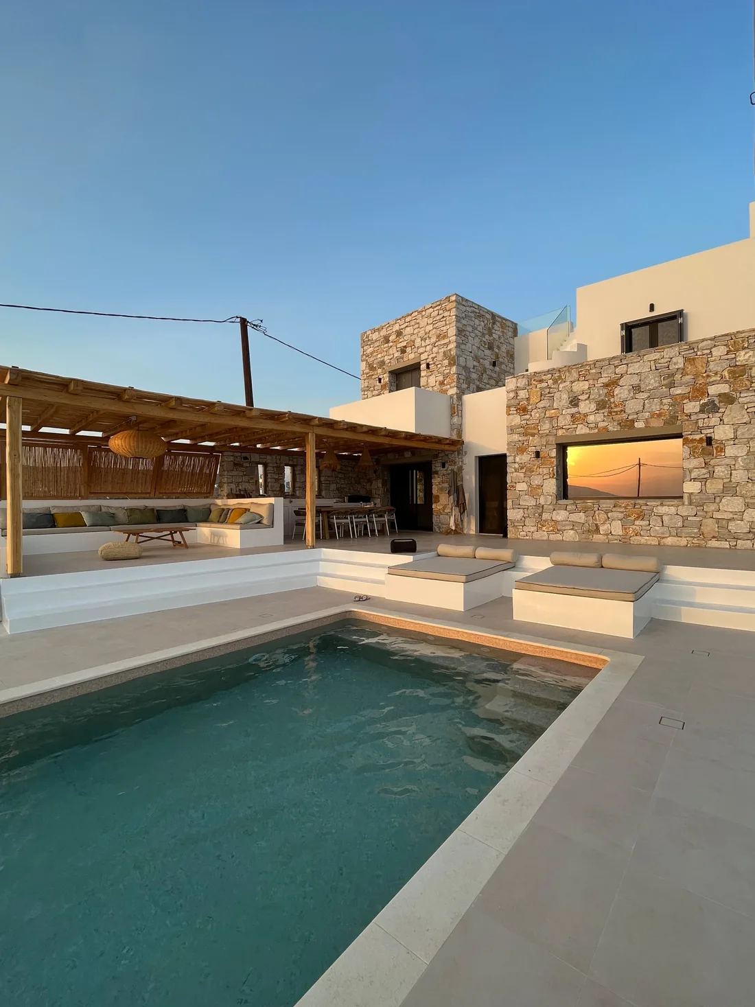 La piscine au crépuscule : bassin turquoise, daybeds maçonnés à matelas écrus, murs de pierre sèche de la villa et coucher de soleil reflété dans la fenêtre