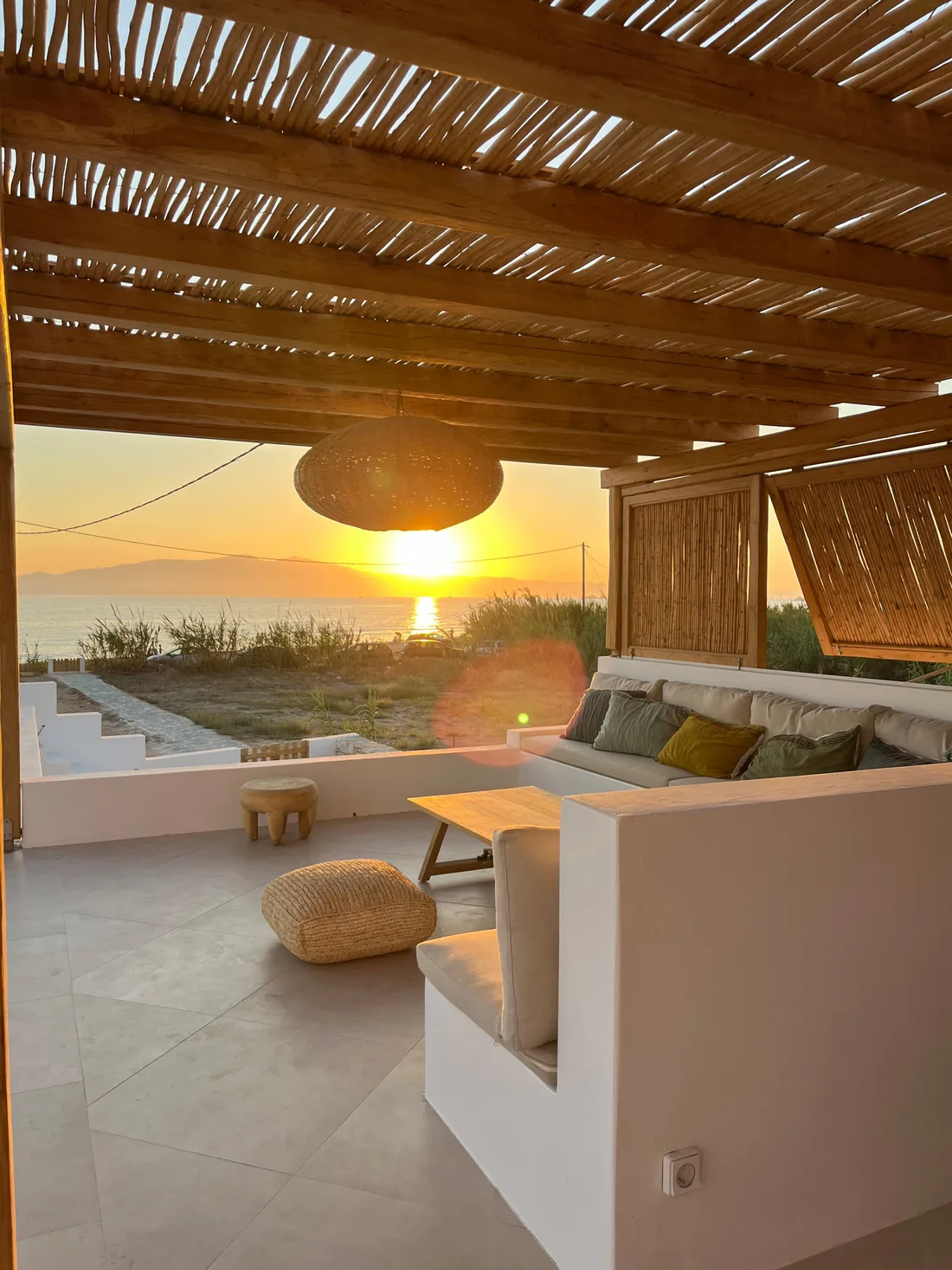 La terrasse couverte au coucher du soleil : pergola de bois et canisse, banquettes maçonnées blanches, suspension en rotin et soleil descendant sur la mer à l'horizon